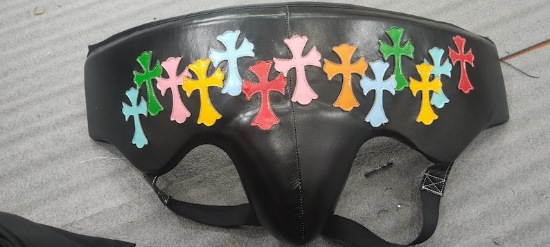 May include: Black leather protective cup with colorful cross appliques. The crosses are in various colors, including green, pink, yellow, and blue. The cup has a contoured shape and adjustable straps.