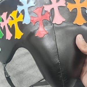 May include: Black leather bag with colorful cross appliques in green, yellow, blue, red, and pink. The bag has a curved shape and a strap. The crosses are outlined in a contrasting color.