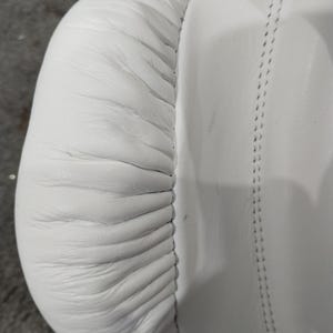 Pu&ograve; includere: Primo piano di un guantone da boxe in pelle bianca. Il guanto presenta cuciture dettagliate e un design imbottito. La pelle ha una consistenza liscia con rughe e pieghe visibili. L'attenzione &egrave; focalizzata sull'area arrotondata e imbottita e sulle cuciture.