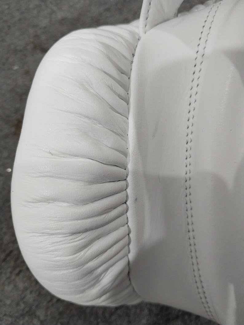 Pu&ograve; includere: Primo piano di un guantone da boxe in pelle bianca. Il guanto presenta cuciture dettagliate e un design imbottito. La pelle ha una consistenza liscia con rughe e pieghe visibili. L'attenzione &egrave; focalizzata sull'area arrotondata e imbottita e sulle cuciture.