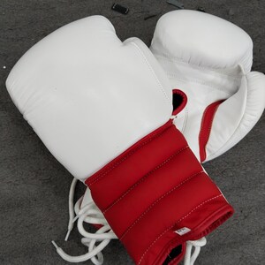 Pu&ograve; includere: Un paio di guantoni da boxe bianchi con dettagli rossi sul polso e sul polsino. I guanti sono in pelle e hanno lacci bianchi. Questi guanti sono progettati per la boxe e altri sport da combattimento.