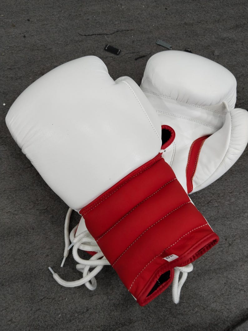 Pu&ograve; includere: Un paio di guantoni da boxe bianchi con dettagli rossi sul polso e sul polsino. I guanti sono in pelle e hanno lacci bianchi. Questi guanti sono progettati per la boxe e altri sport da combattimento.
