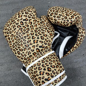 Puede incluir: Un par de guantes de boxeo con estampado de leopardo en marr&oacute;n, negro y beige. Los guantes tienen detalles blancos y palmas de cuero negro. Las correas de mu&ntilde;eca son blancas con ribetes negros. Los guantes son de 16 oz.