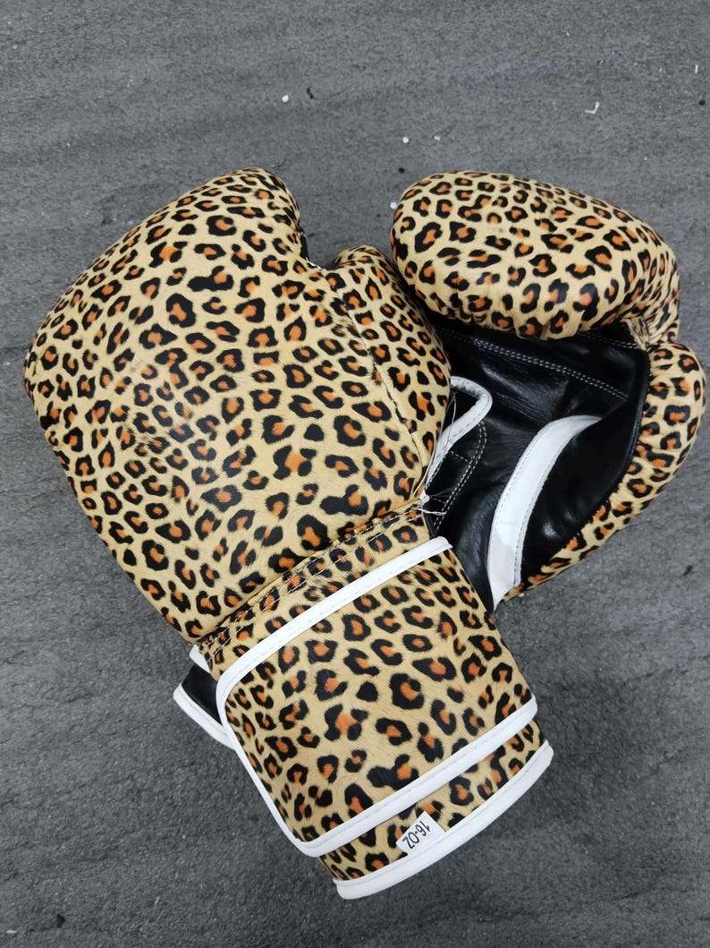 Puede incluir: Un par de guantes de boxeo con estampado de leopardo en marr&oacute;n, negro y beige. Los guantes tienen detalles blancos y palmas de cuero negro. Las correas de mu&ntilde;eca son blancas con ribetes negros. Los guantes son de 16 oz.