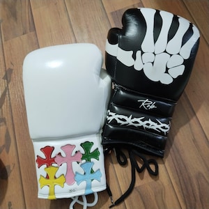 May include: Pair of boxing gloves. One white glove with colorful cross design on the wrist. The other glove is black with a white skeleton hand design and a white thorn design on the wrist.