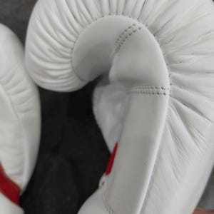 Pu&ograve; includere: Primo piano di guantoni da boxe bianchi con dettagli rossi. I guantoni sono realizzati in un materiale liscio e presentano cuciture dettagliate. Il design si concentra sulla forma curva dei guantoni.