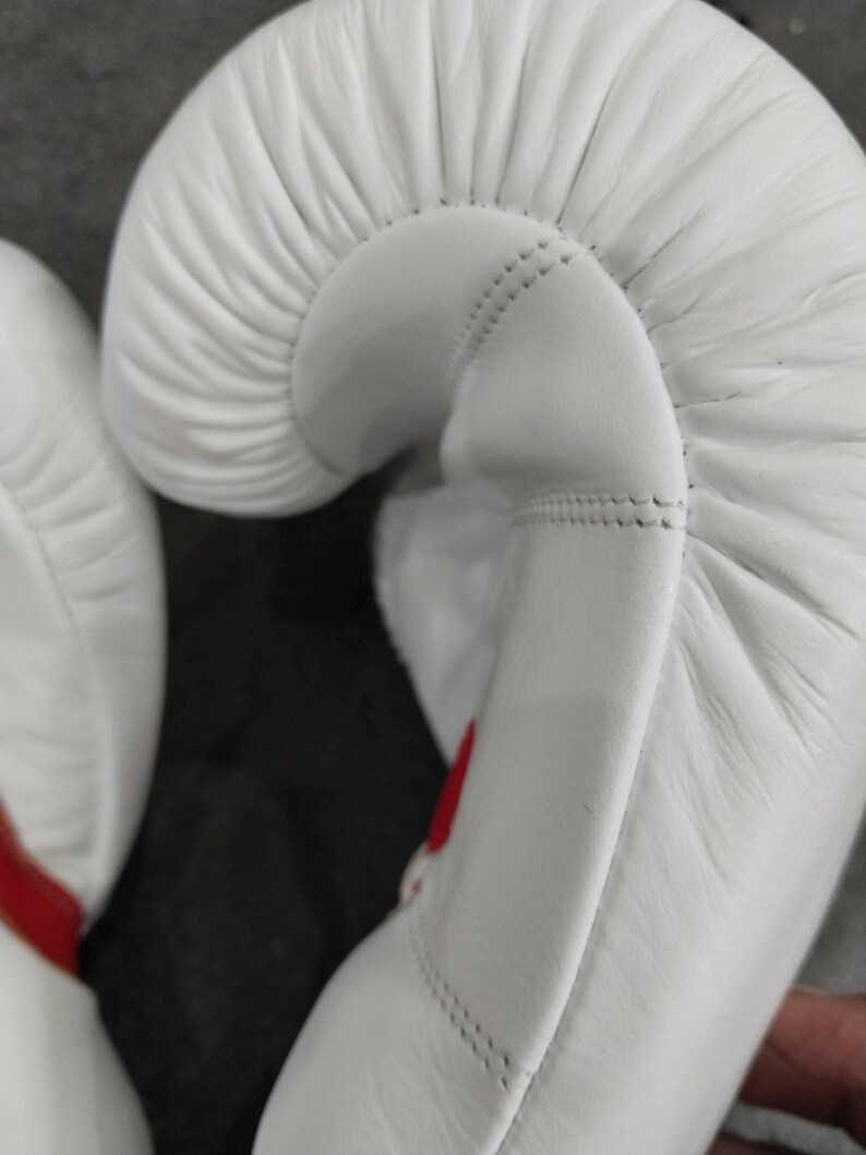 Pu&ograve; includere: Primo piano di guantoni da boxe bianchi con dettagli rossi. I guantoni sono realizzati in un materiale liscio e presentano cuciture dettagliate. Il design si concentra sulla forma curva dei guantoni.