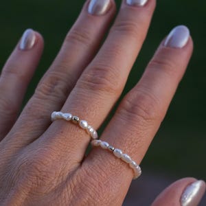 May include: Two delicate pearl rings with gold accents. The rings are worn on the index and middle fingers of a hand.