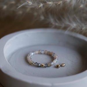 Op de afbeelding: Een kralenarmband met neutrale stenen en goudkleurige accenten ligt op een grijze, cirkelvormige schaal. Twee extra gouden kralen liggen naast de armband. De armband is een sieraad.