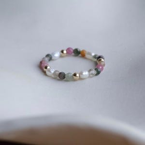 Op de afbeelding: Een kralenring met een mix van kleurrijke edelstenen en witte parels. De ring bevat roze, groene en oranje stenen, afgewisseld met kleine gouden kralen en witte parels.