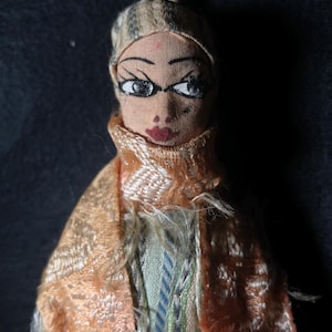 May include: A vintage doll with a painted face, wearing a brown and white hat and a peach-coloured scarf. The doll is dressed in a multi-coloured patterned fabric.