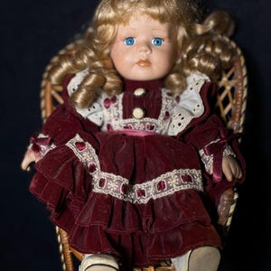 Puede incluir: Una muñeca de porcelana con cabello rubio rizado y ojos azules, vestida con un vestido de terciopelo burdeos con ribete de encaje, sentada en una silla de mimbre.