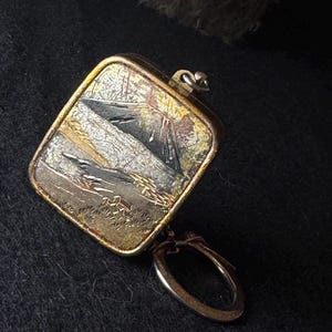 Puede incluir: Un colgante de metal de forma cuadrada que presenta una escena de paisaje detallada con una montaña, árboles y tonos texturizados en oro y plata. El colgante tiene un bucle de metal redondeado en la parte inferior y una pequeña anilla en la parte superior.