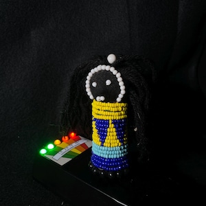 May include: A small beaded doll with black yarn hair, a black face with white bead eyes, and a colourful beaded dress with yellow, blue, and white beads.