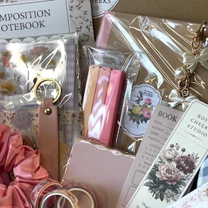 May include: A stationery gift set containing a composition notebook, a pink scrunchie, a leather keychain, three pastel highlighters, a floral bookmark labeled Rose Cheek Studio, a book tracker card, and decorative washi tape rolls in clear packaging.