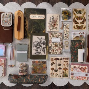 May include: An assortment of stationery items, including notebooks, pens, stickers, and a pencil case, arranged on a white scalloped surface. The items feature floral and botanical designs, with a sketchbook prominently displayed. Colors include brown, green, and pastel hues.