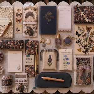 May include: An assortment of stationery and accessories, including notebooks, pens, a pencil case, and a pouch, arranged on a scalloped surface. Floral and butterfly motifs are prominent, with a color palette of brown, cream, and navy.