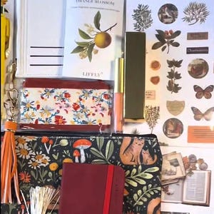 May include: An assortment of stationery items, including a floral print pouch, a red notebook, and decorative stickers. A lip gloss and a card with an orange blossom illustration are also visible. The items are arranged on a surface, creating a visually appealing collection.