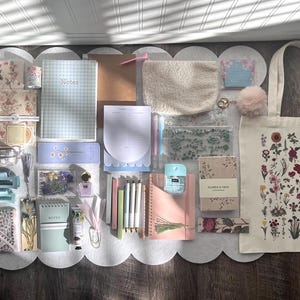 May include: A collection of stationery items, including notebooks, pens, and a tote bag. The tote bag features a floral design. Other items include a pencil case, scrunchies, and a small pouch. The color palette is soft, with pastel pinks, blues, and greens.