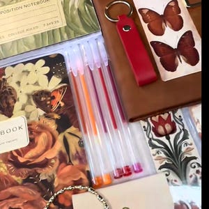 May include: A collection of stationery items including a floral-patterned notebook, a red leather keychain, and a pack of colorful pens. The pens have orange, red, and pink ink. The notebook has a floral design with butterflies.
