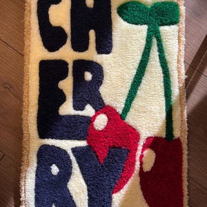 Op de afbeelding: Een rechthoekig, cr&egrave;mekleurig vloerkleed met het woord "CHERRY" in marineblauwe letters. Twee rode kersen met groene stelen en bladeren staan rechts. Het vloerkleed heeft een getextureerde pool en een beige rand.