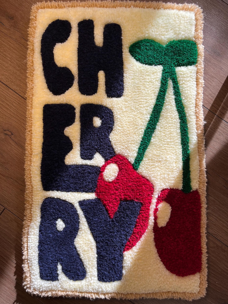 Op de afbeelding: Een rechthoekig, cr&egrave;mekleurig vloerkleed met het woord "CHERRY" in marineblauwe letters. Twee rode kersen met groene stelen en bladeren staan rechts. Het vloerkleed heeft een getextureerde pool en een beige rand.