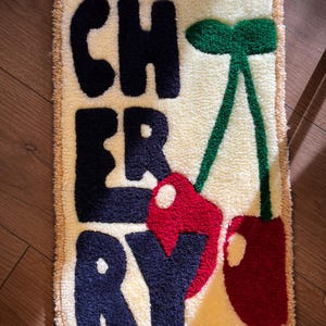 Op de afbeelding: Een rechthoekig tapijt met het woord "CHERRY" in marineblauwe letters. Het tapijt heeft een cr&egrave;mekleurige achtergrond met een beige rand en een groene stengel met twee rode kersen.