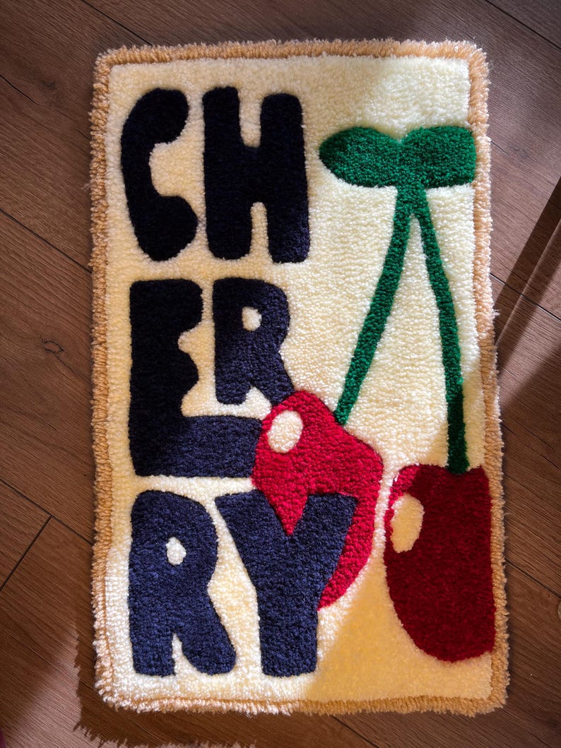 Op de afbeelding: Een rechthoekig tapijt met het woord "CHERRY" in marineblauwe letters. Het tapijt heeft een cr&egrave;mekleurige achtergrond met een beige rand en een groene stengel met twee rode kersen.
