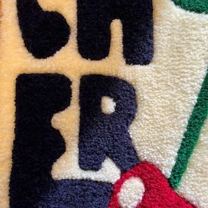 Op de afbeelding: Close-up van een getextureerd tapijt met het woord "CHER" in marineblauw, rood en groen tegen een cr&egrave;me- en gele achtergrond. De letters zijn dik en zacht, waardoor een tactiele uitstraling ontstaat. Het tapijt heeft een handgemaakte uitstraling.