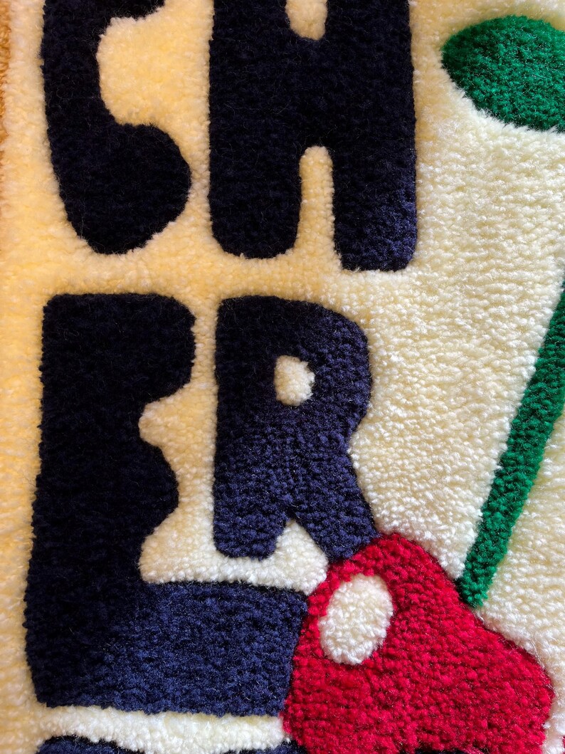 Op de afbeelding: Close-up van een getextureerd tapijt met het woord "CHER" in marineblauw, rood en groen tegen een cr&egrave;me- en gele achtergrond. De letters zijn dik en zacht, waardoor een tactiele uitstraling ontstaat. Het tapijt heeft een handgemaakte uitstraling.