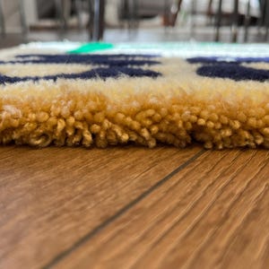 Op de afbeelding: Close-up van een kleurrijk, getextureerd tapijt. Het tapijt heeft een gele rand met marineblauwe, rode en groene accenten. Het tapijt is gemaakt van een zacht, gelust materiaal.