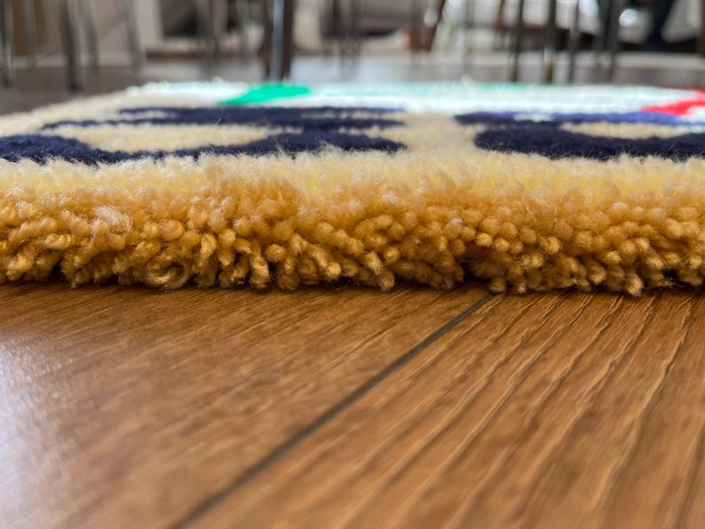 Op de afbeelding: Close-up van een kleurrijk, getextureerd tapijt. Het tapijt heeft een gele rand met marineblauwe, rode en groene accenten. Het tapijt is gemaakt van een zacht, gelust materiaal.