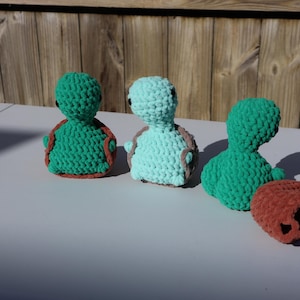 May include: Three crocheted turtle toys. One is green, one is light blue, and one is dark green. The turtles are all sitting on a white surface.