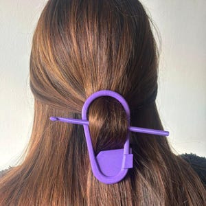 May include: A purple hair clip in the shape of a safety pin secures a section of brown hair. The clip is made of a solid, smooth material and is a vibrant shade of purple. The hair is long and straight.