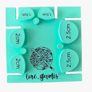 Può includere: Uno strumento quadrato in plastica verde menta per misurare pezzi all'uncinetto, con sei ritagli a forma di goccia etichettati 1,5 cm, 2 cm e 2,5 cm. Il centro mostra un grafico di un gomitolo di lana con un uncinetto e il testo lore_gumis.