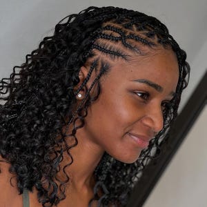 May include: A woman with black hair styled in small braids with curly ends. The braids are parted on the side and styled in a pattern on the top of her head.