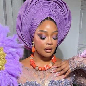 May include: A woman wearing a large, purple, pleated headwrap, a coral necklace, and coral earrings. She is wearing a sparkly, light purple dress.