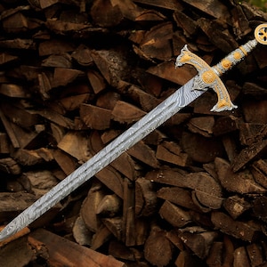 May include: A sword with a double-edged blade featuring a distinct wavy Damascus steel pattern. The hilt is crafted with ornate gold and silver-toned metalwork, including a crossguard and a circular pommel with a central cross emblem.