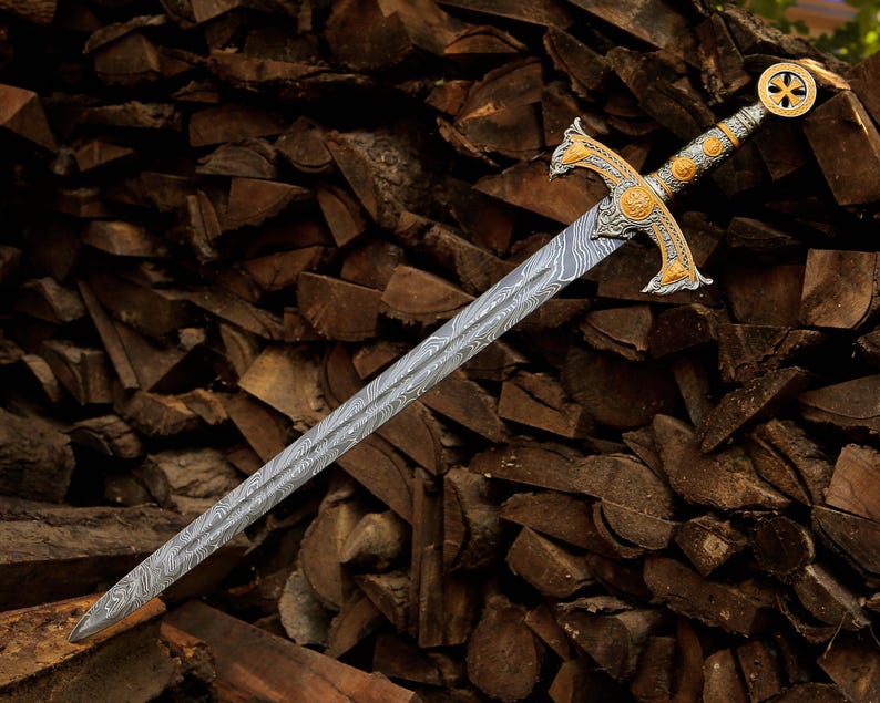 May include: A sword with a double-edged blade featuring a distinct wavy Damascus steel pattern. The hilt is crafted with ornate gold and silver-toned metalwork, including a crossguard and a circular pommel with a central cross emblem.