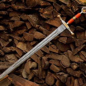 May include: A sword with a long, double-edged blade featuring a distinct wavy Damascus steel pattern. The hilt includes a gold-toned crossguard with ornate engravings, a bright orange grip, and a circular pommel containing a gold-colored emblem.