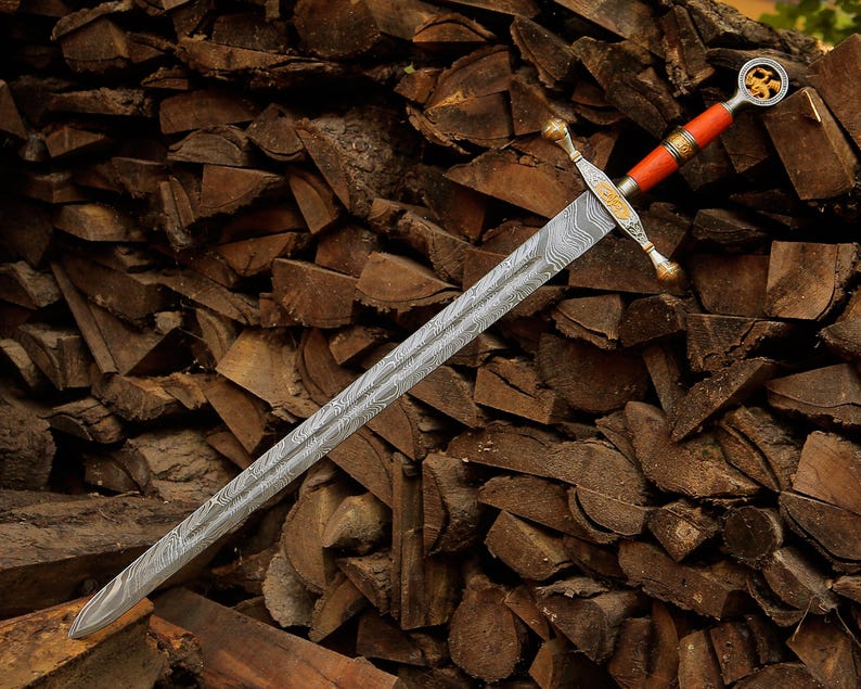 May include: A sword with a long, double-edged blade featuring a distinct wavy Damascus steel pattern. The hilt includes a gold-toned crossguard with ornate engravings, a bright orange grip, and a circular pommel containing a gold-colored emblem.