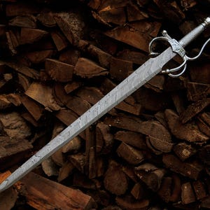 May include: A sword with a double-edged blade featuring a distinct wavy Damascus steel pattern. The hilt includes a polished silver-colored metal crossguard with curved quillons and a textured grip, terminating in a rounded pommel at the end.