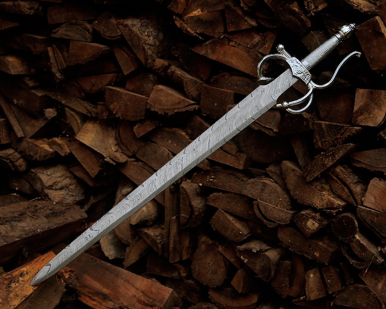 May include: A sword with a double-edged blade featuring a distinct wavy Damascus steel pattern. The hilt includes a polished silver-colored metal crossguard with curved quillons and a textured grip, terminating in a rounded pommel at the end.