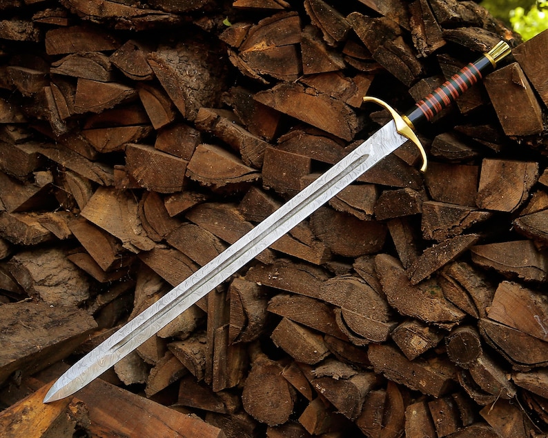 May include: A double-edged sword with a patterned steel blade, a gold-colored crossguard, and a black handle wrapped with thin red bands. The pommel is gold-colored, and the blade features a central fuller running down the length of the metal.