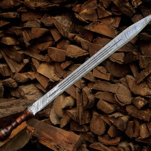 May include: A double-edged sword featuring a patterned Damascus steel blade with a central fuller and a dark brown wooden handle with a rounded pommel. The blade displays a distinct wavy, layered metal texture throughout its length.