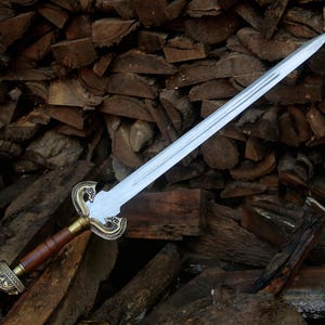 May include: A double-edged steel sword featuring a central fuller, a brown leather-wrapped grip, and an ornate brass-toned crossguard and pommel with intricate engraved detailing. The blade is straight and tapers to a sharp, pointed tip.