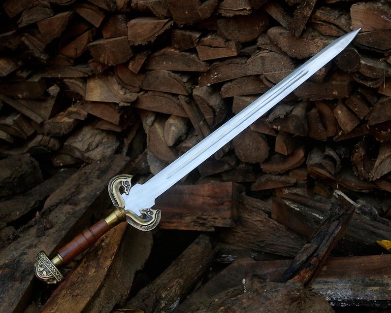 May include: A double-edged steel sword featuring a central fuller, a brown leather-wrapped grip, and an ornate brass-toned crossguard and pommel with intricate engraved detailing. The blade is straight and tapers to a sharp, pointed tip.