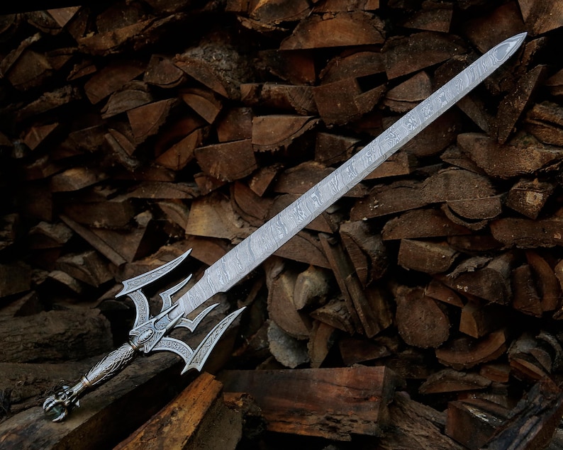 May include: A silver-toned sword featuring a long, double-edged blade with a visible layered metal pattern. The hilt displays intricate engravings, a complex crossguard with multiple pointed prongs, and a textured handle with a decorative pommel.