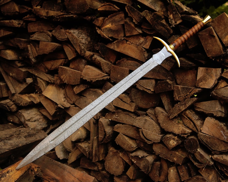 May include: A double-edged sword with a patterned Damascus steel blade, a gold-colored metal crossguard, and a brown leather-wrapped handle with a gold-colored pommel. The blade features a distinct wavy, layered metal texture throughout its length.