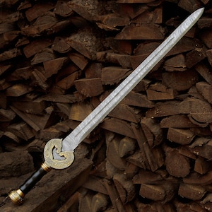 May include: An intricately designed sword with a patterned blade and a decorative handle. The handle has a black grip, gold detailing, and a brass-coloured guard with a horse motif. The sword is displayed against a backdrop of stacked firewood.